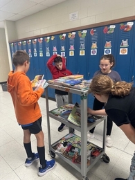 Four students in a school hallway gather around a rolling cart filled with books to choose their free prizes for Family Reading Week.