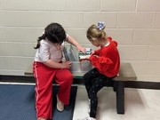 Two young girls sit side-by-side on a bench in front of a cinderblock wall. One student in a white shirt and red pants leans over a plastic bin filled with books, while the other student in a red sweater and heart-patterned leggings helps her flip through