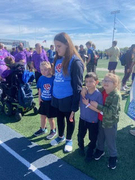 Four young students, two in blue "HEAT" shirts and two in casual wear, stand closely together on artificial turf next to a blue running track. They are looking down or off-camera, surrounded by other attendees, including someone in a motorized wheelchair,