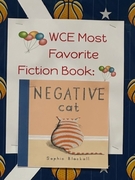 A school bulletin board with a basketball-themed background. A white sign with colorful balloon graphics reads "WCE Most Favorite Fiction Book" in red text. Below it is the cover of the book "Negative Cat" by Sophie Blackall, showing an orange-and-white s