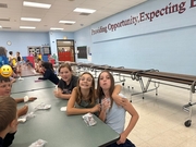 Three elementary students sitting close together at a cafeteria table smiling and posing for the camera with their ice cream wrappers in hand, with a school wall slogan in the background.