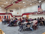 A wide view of the gymnasium showing multiple families, parents, and young children seated at tables on floor tarps. The back wall displays the West Central logo and the school motto: "Providing Opportunity, Expecting Excellence."