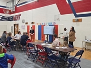 Parents, young students, and family guests seated around folding tables on a gym floor covering, listening to school staff standing next to a large digital display screen. The gymnasium wall features red and white painted accents with "HEAT" written on th