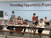 Parents, guests, and young children seated together at a blue cafeteria table with paper handouts, pencils, and activities spread out in front of them. In the background, other families participate beneath a wall banner reading "Providing Opportunity, Exp